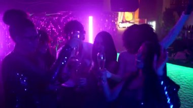 Mor neon ışıklı bir gece kulübünde dans eden ve kadehlerini şampanya bardaklarıyla kaldıran neşeli bir grup genç arkadaş birlikte kutluyor ve eğleniyorlar.