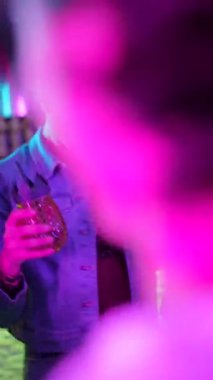 Kokteyllerle dans eden ve bir gece kulübünde kutlama yapan genç çok ırklı arkadaşlar, parlak parlak bir perde arkası olan parlak mor ve mavi neon ışıkların altında eğleniyorlar.