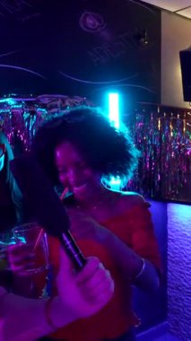 Genç çoklu etnik arkadaşlar dans ediyor, mikrofonla şarkı söylüyor ve renkli neon ışıkları olan canlı bir gece kulübünde kokteyller içiyor. Saf eğlence ve gece hayatı eğlencesi anları yakalıyorlar.