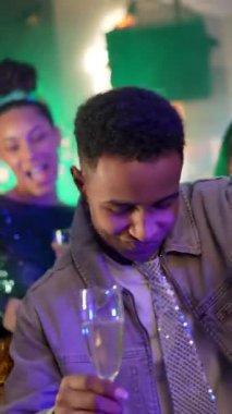 Parlak kravatlı, gece kulübünde dans eden ve şampanya içen mutlu genç siyahi adam. Renkli neon ışıklarıyla neşeli bir atmosfer, gülümsüyor ve arkadaşlarıyla müziğin tadını çıkarıyor.