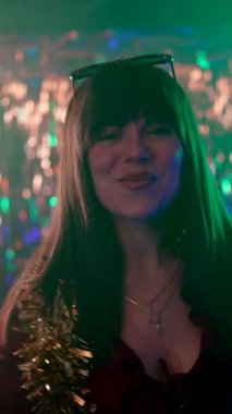 Uzun siyah saçlı neşeli genç bir kadın omuzlarında süslü bir çelenkle dans ediyor ve gülümsüyor. Gece kulübü partisinde elinde renkli ışıkları olan bir bardak şampanya tutarken.