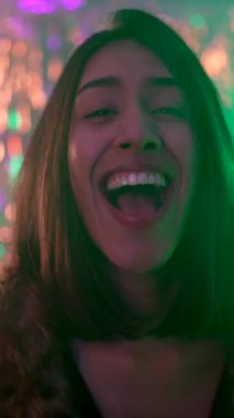 Güzel bir kadın bir bardak şampanyayla dans ediyor, gülüyor ve arka planda renkli ışıklar olan bir partide eğleniyor, özel bir olayı neşe ve heyecanla kutluyor.