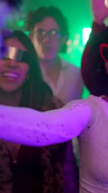 Moda kıyafetleri ve komik güneş gözlükleriyle neşeli çok kültürlü arkadaşlar enerjik bir şekilde dans ediyor ve neon ışıkları ve hareketli bir atmosferi olan bir gece kulübünde parti sırasında birlikte gülüyorlar.