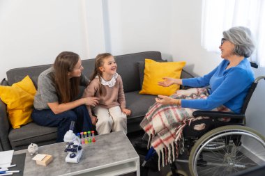 Üç nesil boyunca aile bireyleri sıcak etkileşim içinde, tekerlekli sandalyedeki yaşlı bir kadın, kızı ve torunuyla bir kanepede nişanlanıyor, sevgi dolu ve destekleyici bir aile sahnesi yaratıyor.