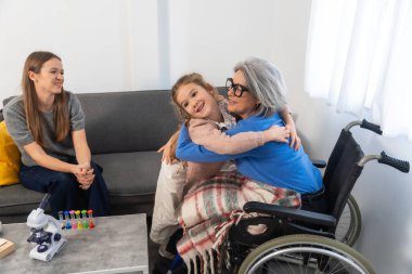 Küçük bir kız tekerlekli sandalyede oturan büyükannesine sarılıyor. Bir kadın da koltuktan izliyor. Nesiller arası bağlantıyı ve aile bakımını gösteriyor.