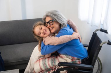 Yaşlı büyükanneyi kucaklayan gri saçlı ve gözlüklü bir torun. Tekerlekli sandalyede oturuyor. Bir ev ziyareti sırasında ya da günlük bakım sırasında şefkat ve nesiller arası bir bağ paylaşmak