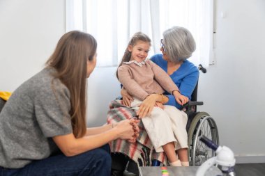 Aile bireyleri, bir ziyaret sırasında gülümseyen bir torunla birlikte, tekerlekli sandalyede büyükannesinin kucağında rahatça oturup, sevgiyi ve nesiller arası bağları sembolize ediyorlar.