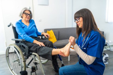 Tekerlekli sandalyede oturan yaşlı bir kadına ayak muayenesi ve fizik tedavi uygulayan profesyonel bir bakıcı, sağlık, rehabilitasyon ve destekli yaşam desteğine odaklanıyor.