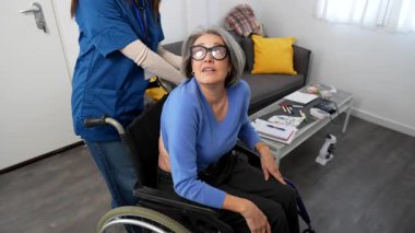 Tekerlekli sandalyedeki yaşlı bir kadın göğüs ağrısı ve nefes darlığı çekerken bir bakıcı bakımevinde stetoskop ile acil tıbbi yardım sağlar.