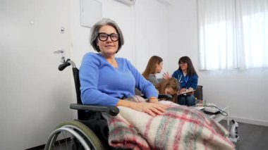 Engelli bir kadın tekerlekli sandalyede otururken gülümsüyor. Bakımevinde battaniyeyle örtülmüş, ailesi ve arka planda sohbet eden bir bakıcı var.