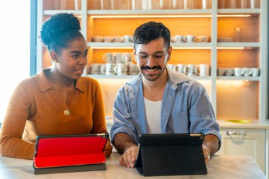 Çeşitli iş arkadaşları, bir mola sırasında dijital tabletlerin içeriğini inceleyerek ve işbirliği içinde çalışarak, ortak çalışan bir mutfak alanında fikirleri tartışıyor.