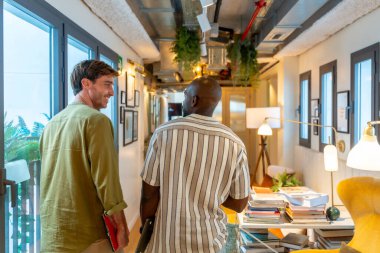 Modern bir ofis veya çalışma alanında yürüyen ve sohbet eden iki farklı meslektaş, mola sırasında fikirleri paylaşıyor ve işbirliği yapıyor, takım çalışması ve profesyonel etkileşimi betimliyor