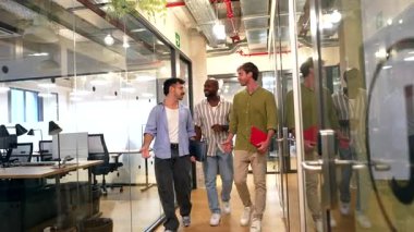 Üç çok kültürlü erkek iş arkadaşı modern cam duvarlı ofis koridorunda birlikte yürüyor, dostane bir sohbete girişiyor ve işten ayrılırken fikirlerini paylaşıyorlar.