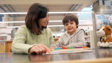 Bakıcı bir anne, genç oğluna halk kütüphanesindeki bir masada renkli bir eğitim oyuncağı öğreterek, erken çocukluk gelişimini, öğrenmeyi ve aile bağlarını oyun oynayarak teşvik ediyor.