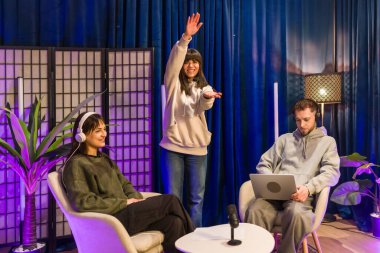 Modern bir ev stüdyosunda işbirliği yapan genç yetişkinler, bir podcast kaydediyor ve sosyal medya ve canlı yayın platformları için mikrofonlar, laptoplar ve renkli ışıklandırmalarla video içeriği oluşturuyorlar.