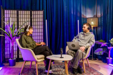 İki genç içerik yaratıcısı, bir erkek ve bir kadın, kulaklık takıyor ve sandalyelerde oturuyorlar, modern bir stüdyo ortamında mikrofon ve dizüstü bilgisayarla ses yayını yapıyorlar.