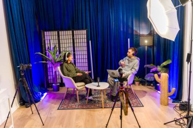 Profesyonel bir stüdyoda sosyal medya podcast 'i kaydeden çeşitli genç insanlar, mikrofonlar, kameralar ve aydınlatma ekipmanlarıyla çevrimiçi platformlar için dijital içerik yaratıyorlar.