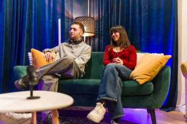 Genç bir adam ve kadın yeşil bir koltukta oturuyor, aktif olarak dinliyor ve ilgileniyorlar. Modern bir stüdyo ortamında, sehpanın üzerinde mikrofon ile bir podcast veya dijital içerik kaydederken.