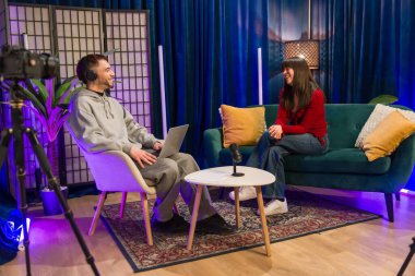 Çeşitli genç içerik yaratıcıları, mikrofon, dizüstü bilgisayar ve kamera ekipmanlarıyla modern bir stüdyoda oturup, bir podcast veya sosyal medya videosu kaydediyor.