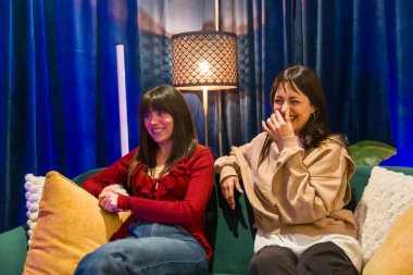 Bir kanepede oturan iki genç kadın, biri gülümsüyor, diğeri ise gülüyor, podcast ve online platformlar için modern bir içerik yaratma stüdyosunda yaratıcı bir şekilde iş birliği yapıyor.