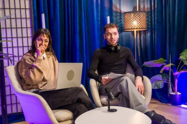 Sosyal medya ve canlı yayın için canlı bir stüdyo ortamında dizüstü bilgisayar, mikrofon ve kulaklık kullanarak bir podcast kayıt oturumuna katılan iki genç içerik yaratıcısı