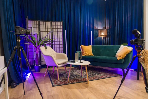 Podcast veya sosyal medya video üretimi için donatılmış modern içerik oluşturma stüdyosu, tripodlardaki kameralar, bir mikrofon, laptop, kanepe, koltuk ve mavi kadife perdeler