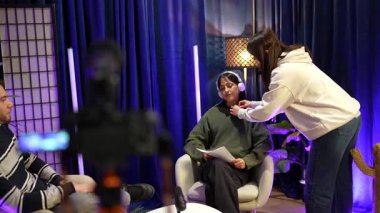 Genç içerik yaratıcıları modern bir podcast stüdyosunda canlı yayına hazırlanıyor. Bir bayan asistan sunucunun gömleğine lavalier bir mikrofon takıyor.