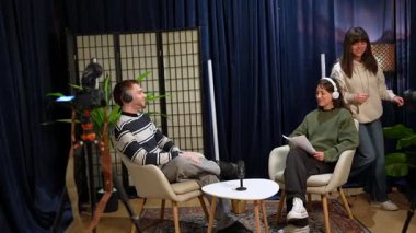 Bir kadın yapımcı yön tarif ederken, iki içerik yaratıcısı bir podcast kaydediyor veya canlı yayın yapıyor. Genç bir adam ve kadın koltuklarda oturmuş mikrofona konuşuyor ve gülümsüyor.