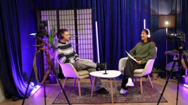 İki içerikli yaratıcı, bir erkek ve bir kadın, kulaklık takıyor ve modern bir stüdyoda profosyonel mikrofonlar, kameralar ve şık mor ışıklandırmalı bir podcast röportajı kaydediyor.