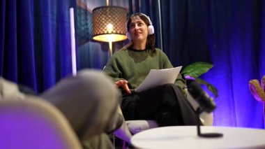 Modern bir podcast stüdyosunda etkileyici bir konuşma sırasında, kulaklık takan ve elinde kağıtlarla konuşan etkileyici genç bir kadın, canlı bir röportaj sırasında puanlarını vurgulamak için el kol hareketi yapıyor.