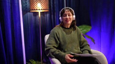 Genç içerik yaratıcısı kulaklık takıyor ve podcast veya canlı yayın yaparken mikrofona konuşuyor, modern profesyonel ışıklandırılmış bir stüdyoda işaret ediyor ve gülümsüyor.