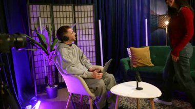 Erkek podcaster kulaklık takıyor. Programına katılan bir bayanı selamlıyor, elini sıkıyor ve modern bir stüdyoda mikrofonlar, kameralar ve profesyonel ışıklandırma ekipmanlarıyla görüşmeye başlıyor.