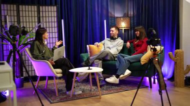 Profesyonel bir stüdyoda kamera ve mikrofonlu bir video yayını yapan üç genç içerik yaratıcısı, sosyal medya seyircileri için canlı bir sohbete girişiyor.