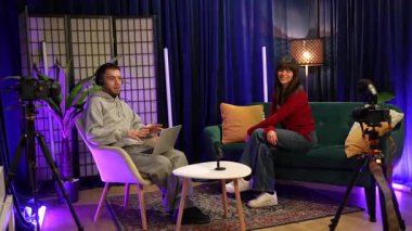 İçerik yaratıcıları, profesyonel bir stüdyoda mikrofonlar ve kameralarla podcast ya da canlı yayın yapıyorlar. Ev sahibi adam, kadını misafir olarak sorgularken birlikte oturup konuşuyorlar.