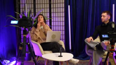 Sosyal medya için içerik yaratan genç bir adam ve kadın, profesyonel bir stüdyoda canlı bir podcast ya da online bir talk show sunuyor mikrofonlar, kameralar ve canlı mor ışıklandırma ile