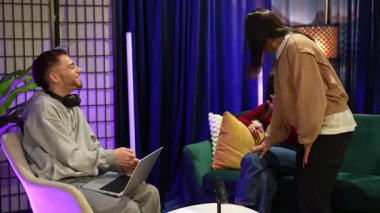 Üç genç içerik yaratıcısı modern bir stüdyoda bir podcast bölümü çekiyor. Erkek sunucu, sosyal medya kanalları için video kaydederken iki kadın konukla görüşüyor.