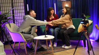 Genç adam bir podcast bölümü kaydediyor. Kameralar, mikrofonlar ve yaratıcı ışıklandırmayla donatılmış modern bir stüdyoda iki neşeli bayan konukla tokalaşıp sohbet ediyor.