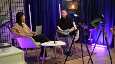 İki genç içerik yaratıcısı, bir erkek ve bir kadın, profesyonel bir stüdyoda kameralar ve mikrofonlarla canlı yayında tartışıyorlar, sosyal medya takipçileri için ilgi çekici içerikler yaratıyorlar.