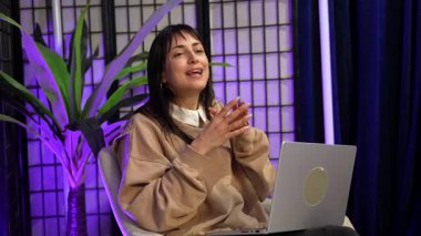 Podcast veya canlı yayın yaparken konuşan ve el kol hareketi yapan kendine güvenen kadın, mor neon ışıkları ve dizüstü bilgisayarlı bir stüdyoda oturuyor ve seyircisine ilgi çekici bir içerik yaratıyor.