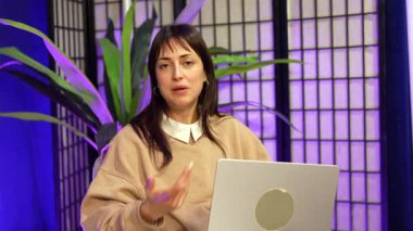 Bir podcast kaydederken ya da canlı yayın yaparken kameraya konuşan ve el kol hareketi yapan kendinden emin genç bir kadın, yaratıcı bir stüdyoda dizüstü bilgisayarı ve canlı mor ışıklandırmasıyla oturuyor.