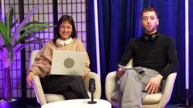 İki genç içerikli yaratıcı, bir erkek ve bir kadın, modern bir stüdyoda canlı bir talk show sunuyorlar, mikrofona konuşuyorlar ve bir sosyal medya yayını için dinleyicileriyle iletişim kuruyorlar.