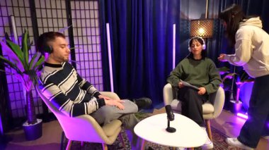 Genç içerik yaratıcıları modern bir stüdyoda neon ışıkları olan bir podcast kaydediyor. Bir erkek ve kadın konuşmacı film çekmeden önce onlara talimat veriyor.