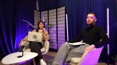 Genç bir adam ve kadın modern bir stüdyoda mikrofonları ve neon ışıkları olan bir talk show, podcast veya vlog kaydediyor, izleyicileri için başlıkları tartışıyor ve çevrimiçi içerik yaratıyorlar.