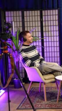 İki genç içerikli yaratıcı, bir erkek ve bir kadın, kulaklık takıyor ve mikrofona konuşuyorlar. Profesyonel bir kayıt stüdyosunda, mor ışıklandırmalı.