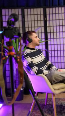 İki genç etkileyici, bir erkek ve bir kadın, kulaklık takıyorlar ve modern bir stüdyoda mor ışıklarla oturuyorlar, sohbet podcastları ya da sosyal medya kanalları için bir röportaj yapıyorlar.