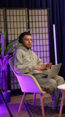 Kulaklıkları ve dizüstü bilgisayarı olan genç bir erkek sunucu canlı yayın için bir kadınla röportaj yapıyor. İkisi de modern ışıklandırması ve profesyonel mikrofonu olan şık bir stüdyoda rahatça oturuyor.