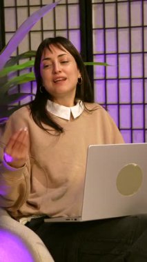 Genç bayan içerik yaratıcısı enerjik bir şekilde konuşur ve işaret eder. Profesyonel bir stüdyoda video yayını ya da video bloğu kaydederken dizlerinin üzerinde mor ortam aydınlatması ve dizüstü bilgisayarı olan bir video bloğu.