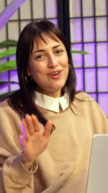 Genç bir bayan içerik yaratıcısı online seyircisi için mor neon ışıkları olan modern bir stüdyoda canlı yayın ya da canlı yayın yaparken konuşur ve işaret eder.