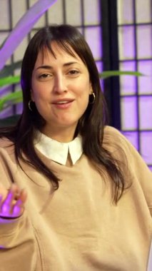 Mor ışıklı yaratıcı bir stüdyoda canlı yayın sırasında konuşan ve el kol hareketi yapan kendinden emin bir kadın vlogger. Sosyal medya veya canlı yayın için video içeriği kaydeden karizmatik bir etkileyiciName