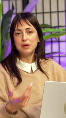 Sosyal medya kanalı için bir podcast ya da canlı yayın yapan genç bir kadın etkileyicisi, mor ortam ışıklandırmasıyla profesyonel bir stüdyoda konuşuyor ve el kol hareketi yapıyor.